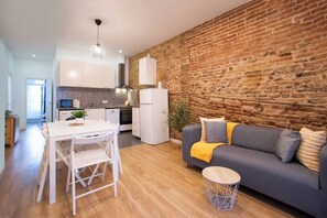 A modern kitchen with white cabinets, a built-in oven, and a microwave. There's a grey sofa with a yellow throw blanket and a small round table with a wireframe base.