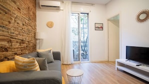 Una sala de estar con pared de ladrillo, un sofá gris, una mesa de centro redonda, un televisor de pantalla plana y un balcón.