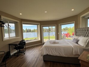 A bedroom with a large bed, a desk, and a chair, with windows offering a view of a lake and houses. - Luxury Lakeview Dream Cottage with Hot Tub (Kearney)