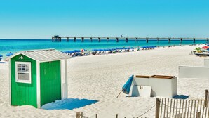 Ein Strand mit einem grünen Gebäude, einer Holzsteg und Strandliegen.