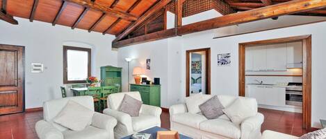 A spacious living area with two white sofas, a coffee table, and a wooden ceiling. A kitchen is visible in the background.