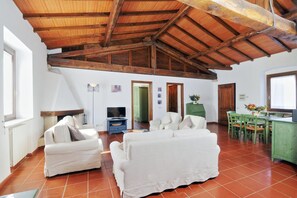 A spacious living area with a high wooden ceiling, white sofas, a television, and a dining area with green chairs.