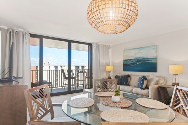 Un soggiorno moderno con un tavolino in vetro, una poltrona e un’area pranzo con sedie. È presente un’ampia finestra vista mare e un balcone.