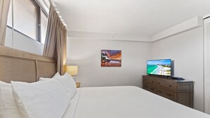 Une chambre d’hôtel avec un grand lit, un téléviseur posé sur une commode en bois, un tableau accroché au mur et une fenêtre avec des rideaux.