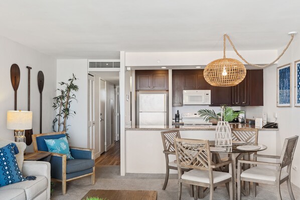 Een moderne keuken met donkere houten kastjes, een witte koelkast en een magnetron. Er is een eethoek met een ronde tafel en stoelen, en een woonkamer met een bank en een lamp.