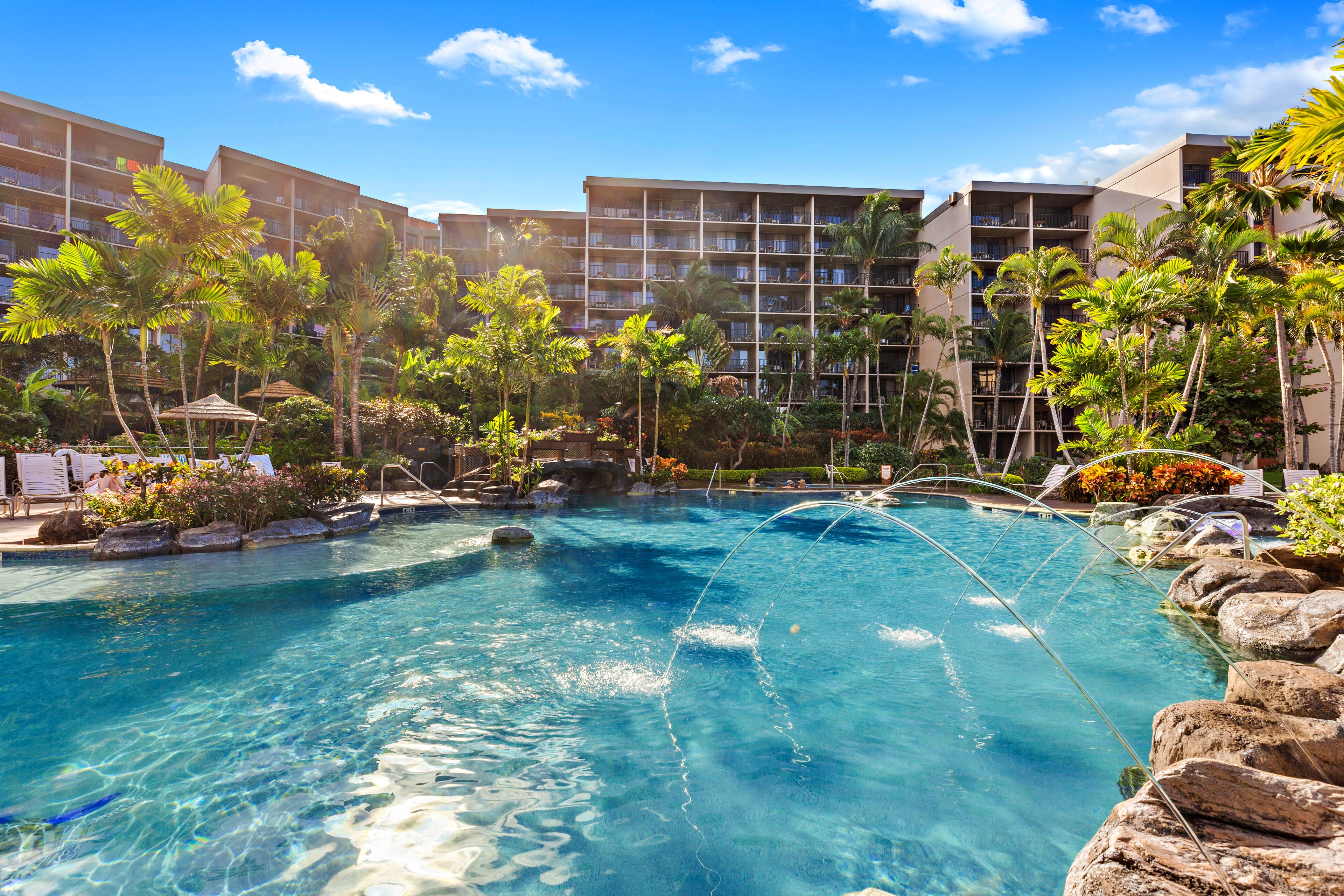 Ein Resort-Pool, umgeben von Palmen und einem mehrstöckigen Gebäude mit Balkonen.