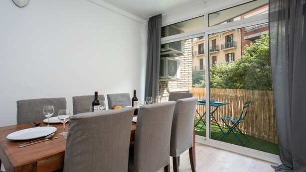 A dining area with a table set for four, a large window with curtains, and a view of a garden and buildings outside. - Sagrada Familia bright apartment with balcony (08013 Barcelona)