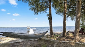 Eine zwischen Bäumen gespannte Hängematte, ein Steg, der ins Wasser führt, und eine Bank unter einem Baum.