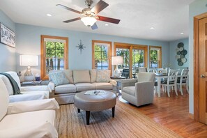 A living room with a ceiling fan, a sofa, a dining table with chairs, and large windows. - Private 100' Waterfront, 4BR - Heidaway Cottage (Sturgeon Bay)
