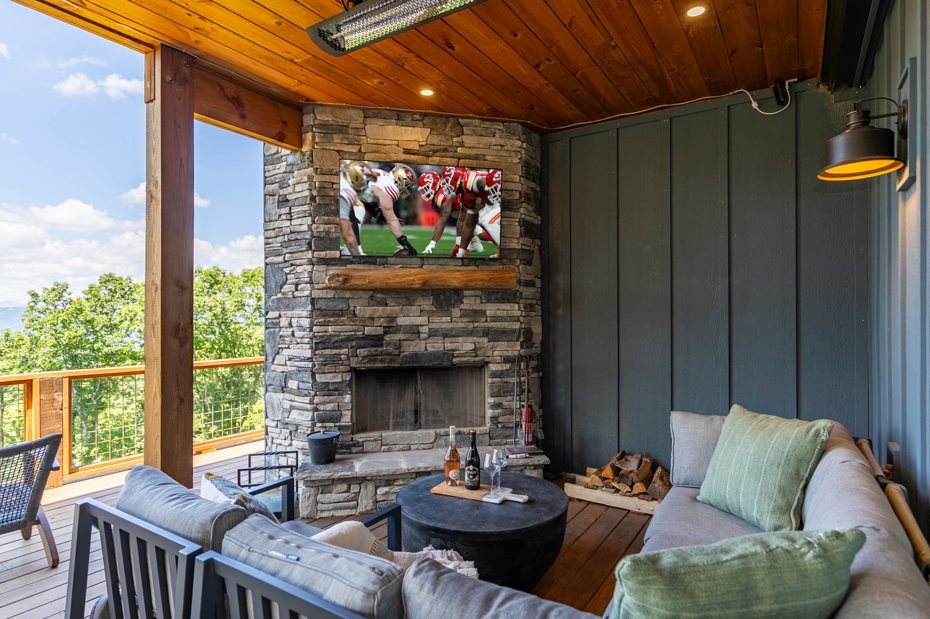 A cozy outdoor seating area with a stone fireplace, a television mounted above it, and a wooden ceiling.