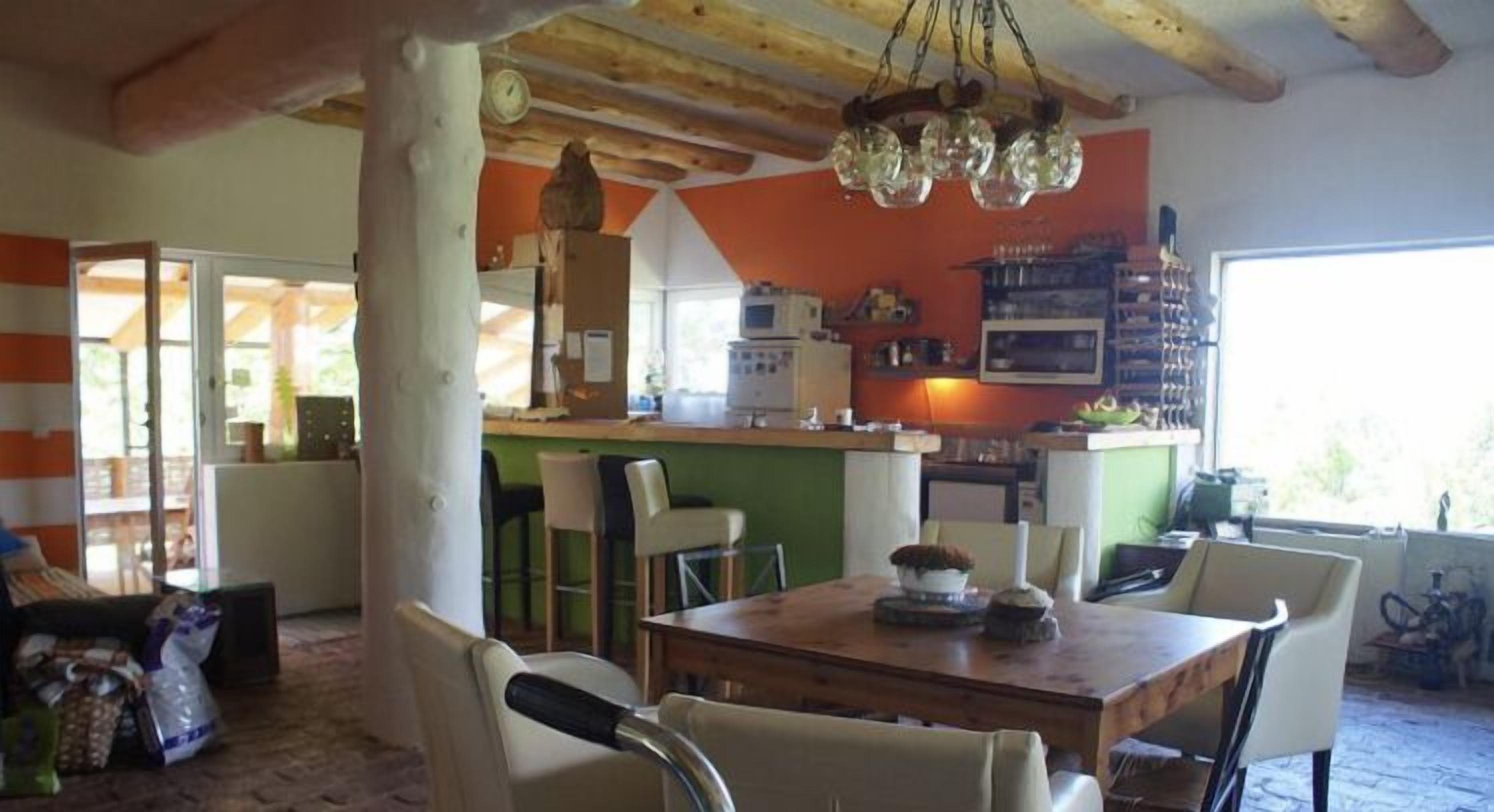 A dining area with a wooden table and chairs, a kitchen in the background, and a window with a view.