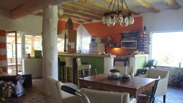 A dining area with a wooden table and chairs, a kitchen in the background, and a window with a view.