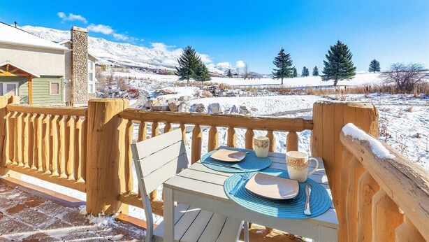 A wooden deck with a table set for two, overlooking a snowy landscape with trees and mountains.