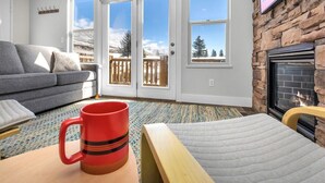 A cozy living room with a grey sofa, a red mug on a wooden table, a fireplace, and a view of snowy mountains through large windows.