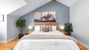 A bedroom with a sloped ceiling, a large bed with a white comforter, two nightstands with lamps, and a mountain landscape painting above the bed.