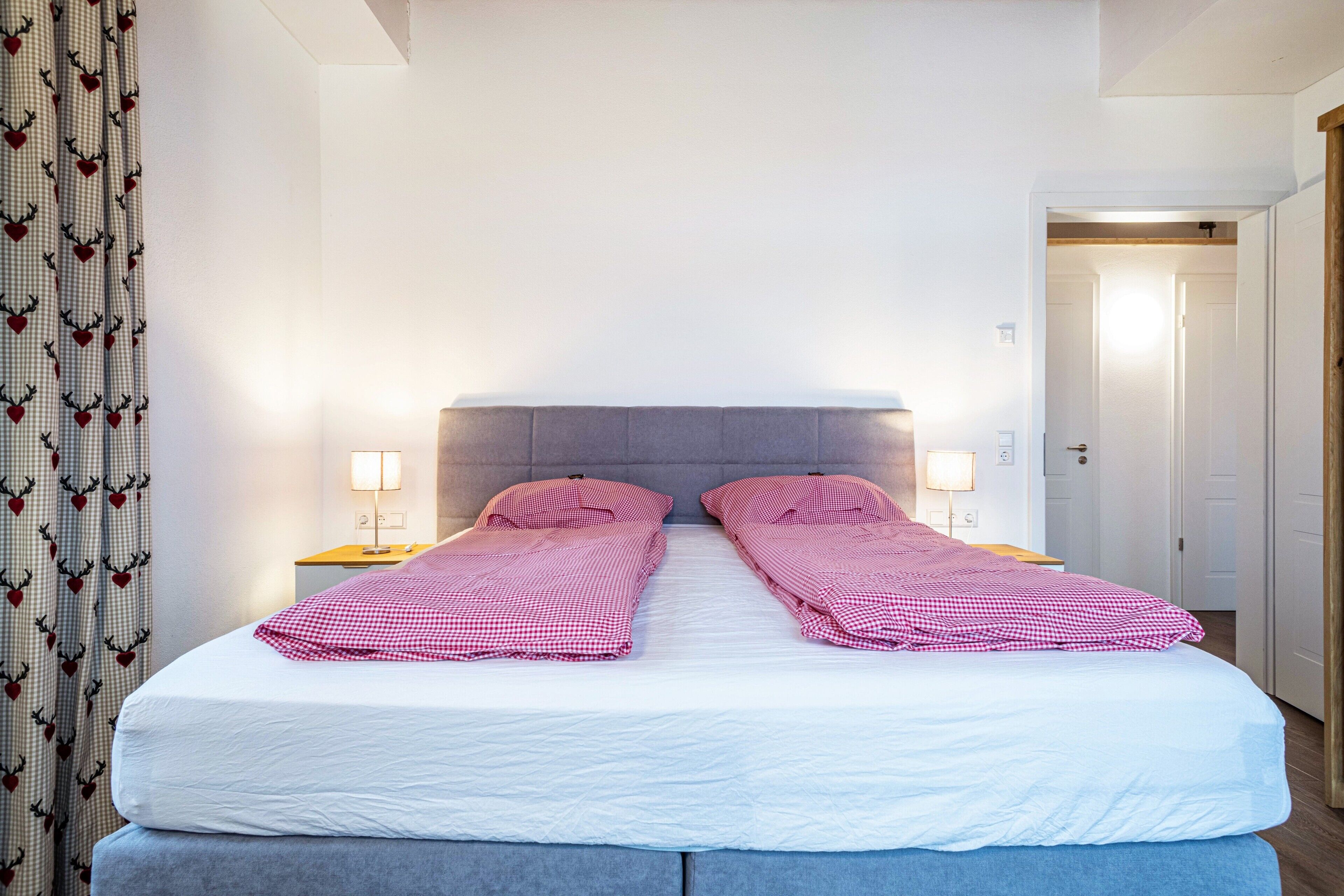 Una cama doble con ropa de cama a cuadros, dos mesitas de noche con lámparas, un cabecero gris y una cortina con estampado de corazones.