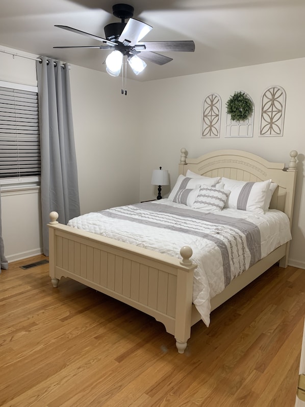 Une chambre à coucher comprenant un lit, un ventilateur de plafond, une fenêtre avec des stores et des œuvres d’art décoratives sur les murs.