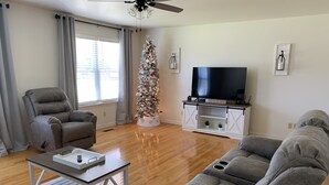 Ein Wohnzimmer mit einem grauen Ecksofa, einem grauen Fernsehsessel, einer weißen Unterhaltungsanlage mit Flachbildfernseher und einem Weihnachtsbaum.