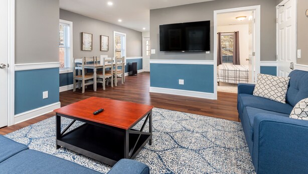 A living room with a TV, blue sofas, a coffee table, and a dining area with chairs and a table.