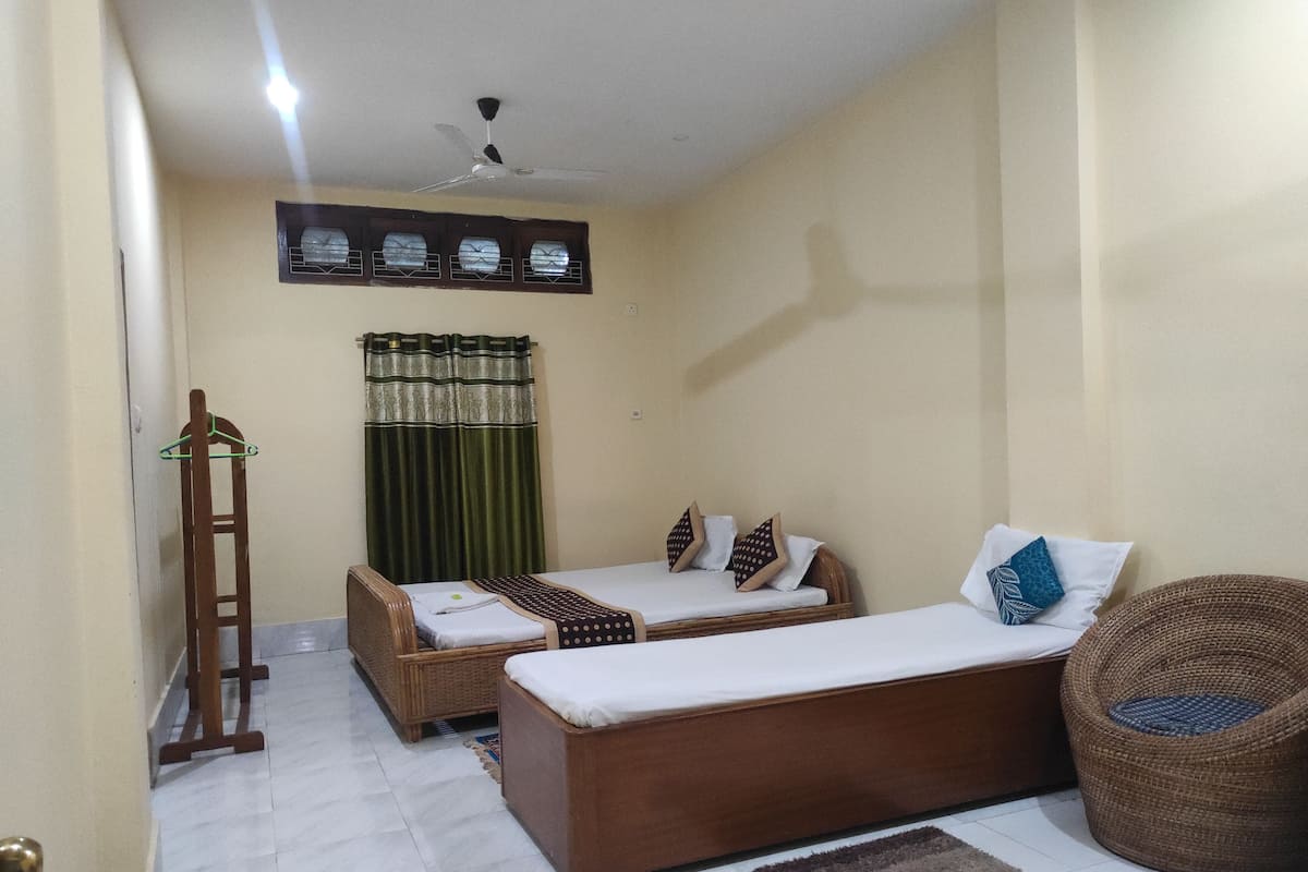 Una habitación con dos camas individuales, un sofá, una silla de mimbre, un ventilador de techo y una ventana con cortinas.
