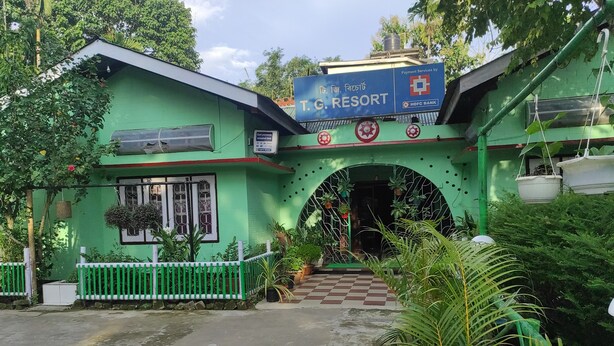 Un edificio verde con un cartel de T.C. Resort y HDFC Bank, una entrada con portón y plantas en macetas.