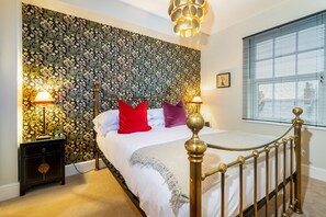 A bedroom with a floral wallpaper, a bed with a gold frame, two bedside tables with lamps, and a window with blinds.