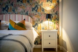 Un dormitorio con cama, almohadas coloridas, una mesita de noche con lámpara y un papel tapiz floral.