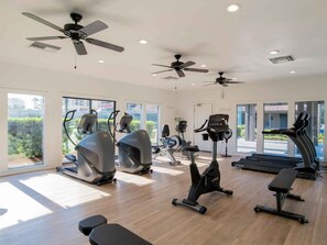 A modern gym with various exercise equipment, large windows, and ceiling fans. - Ground Unit at – Romar House 101A, Easy Beach Access! (Orange Beach)