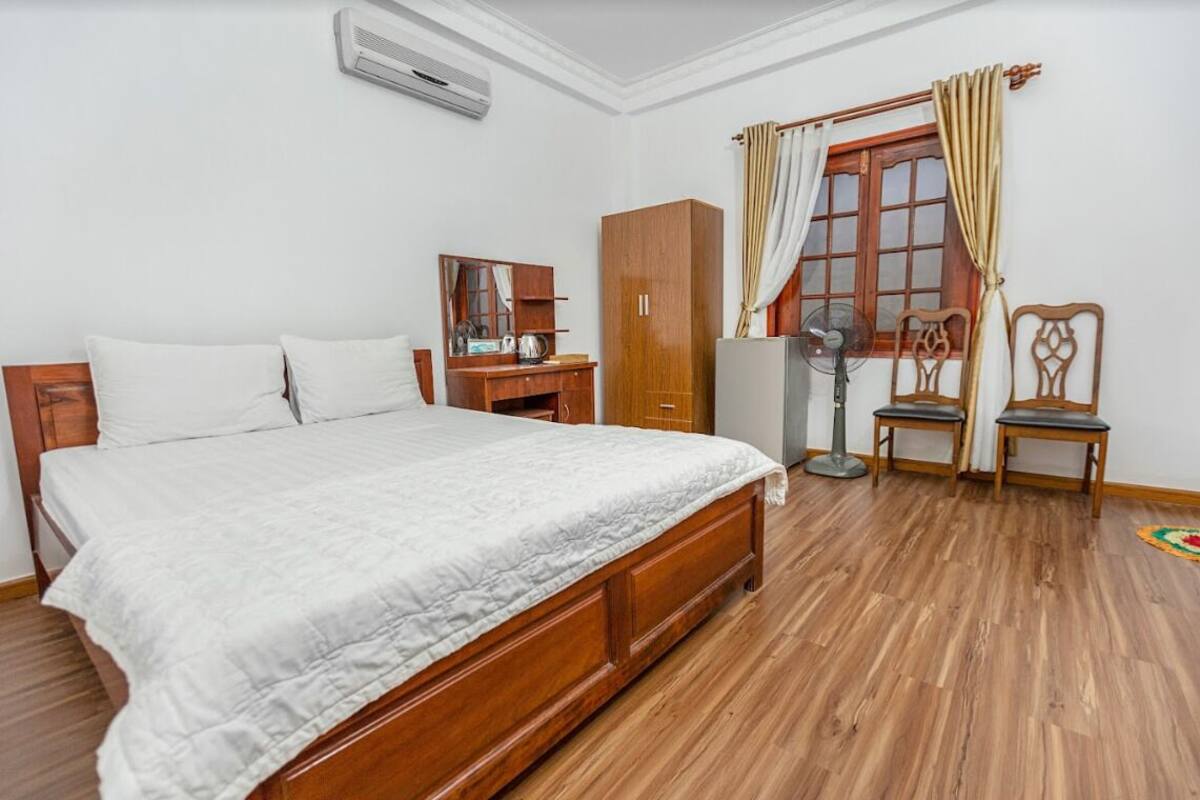 A bedroom with a wooden bed, a wardrobe, a desk with a chair, a standing fan, and a window with curtains.