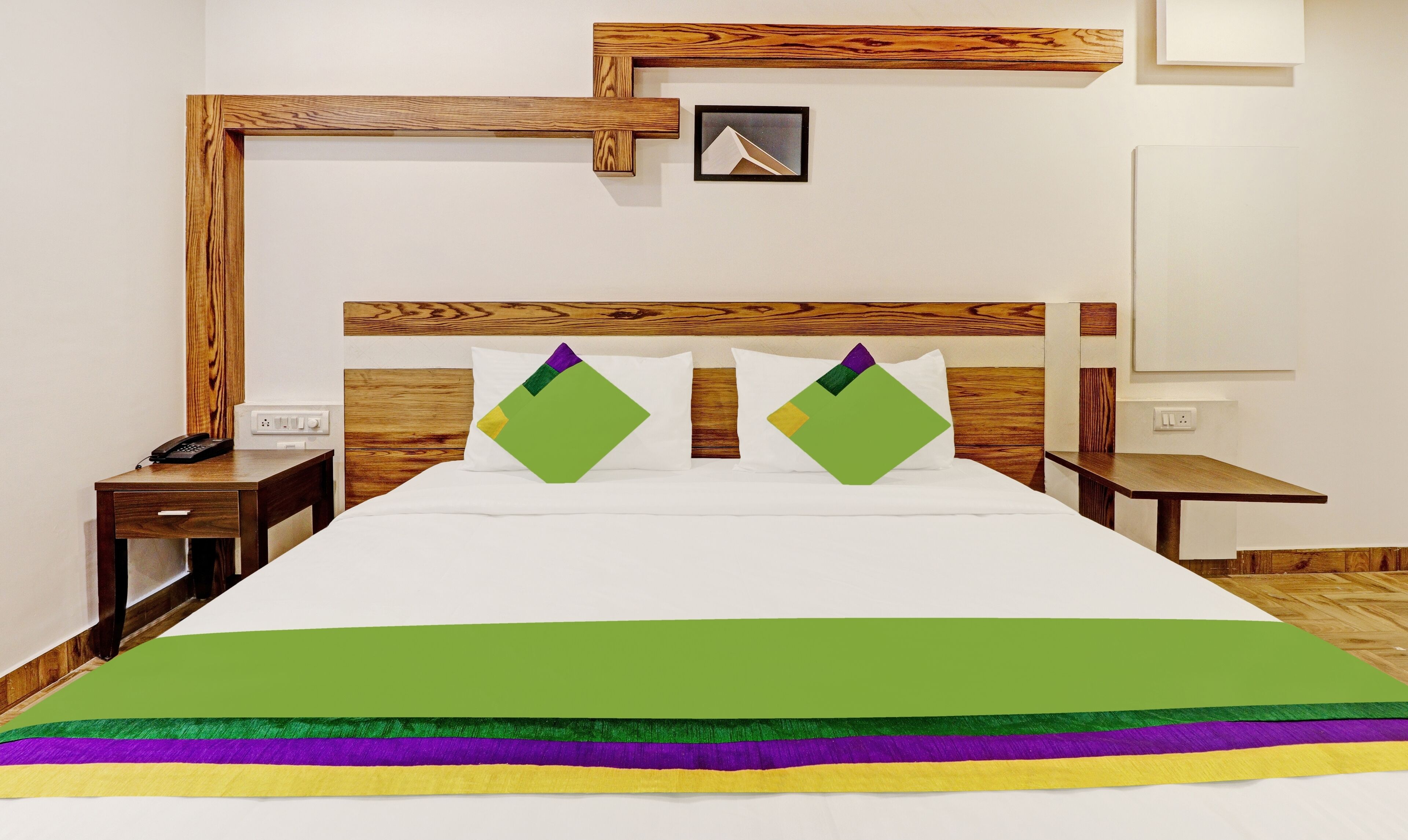 Ein Hotelzimmer mit einem hölzernen Kopfteil, einem Bett mit weißer Bettwäsche, grünen und violetten Kissen sowie einem kleinen Nachttisch mit einem Telefon.