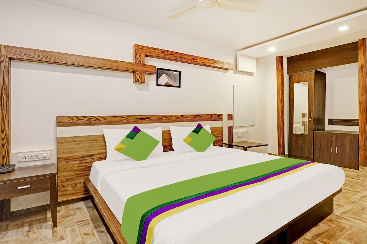 Una habitación de hotel con cabecera de madera, cama con colcha verde y morada, mesita de noche, escritorio y armario.