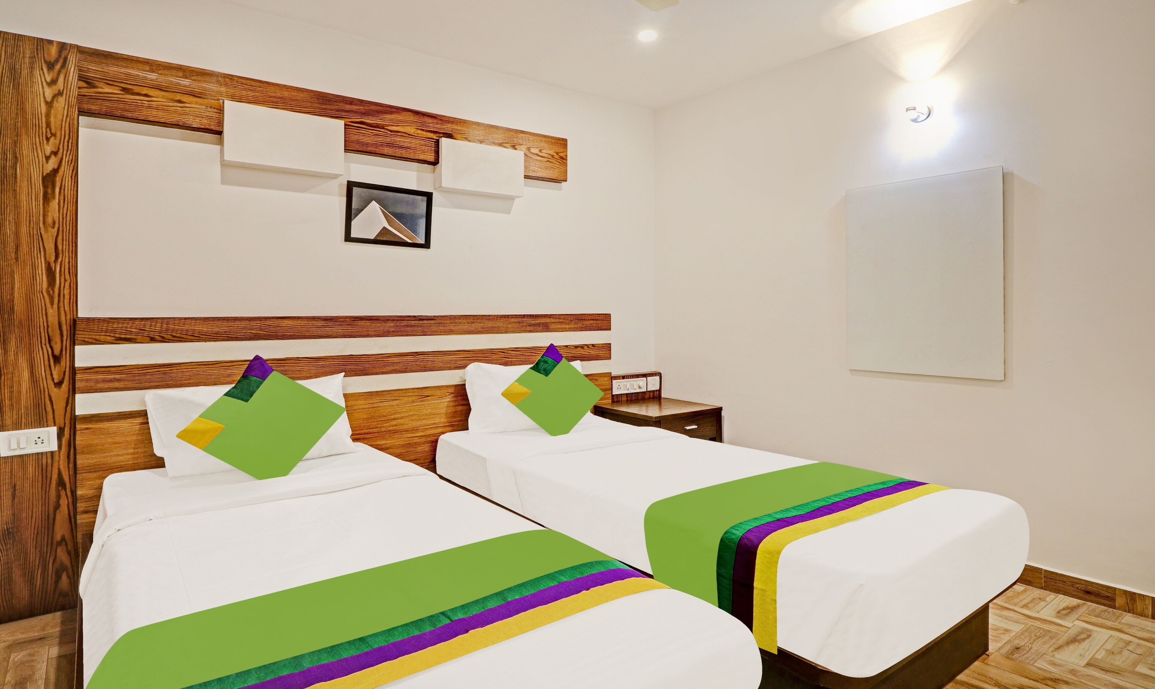 A hotel room with two beds, wooden headboards, a small bedside table, a nightstand, and a wall-mounted light fixture.