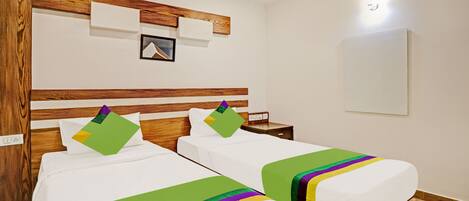 A hotel room with two beds, wooden headboards, a small bedside table, a nightstand, and a wall-mounted light fixture.