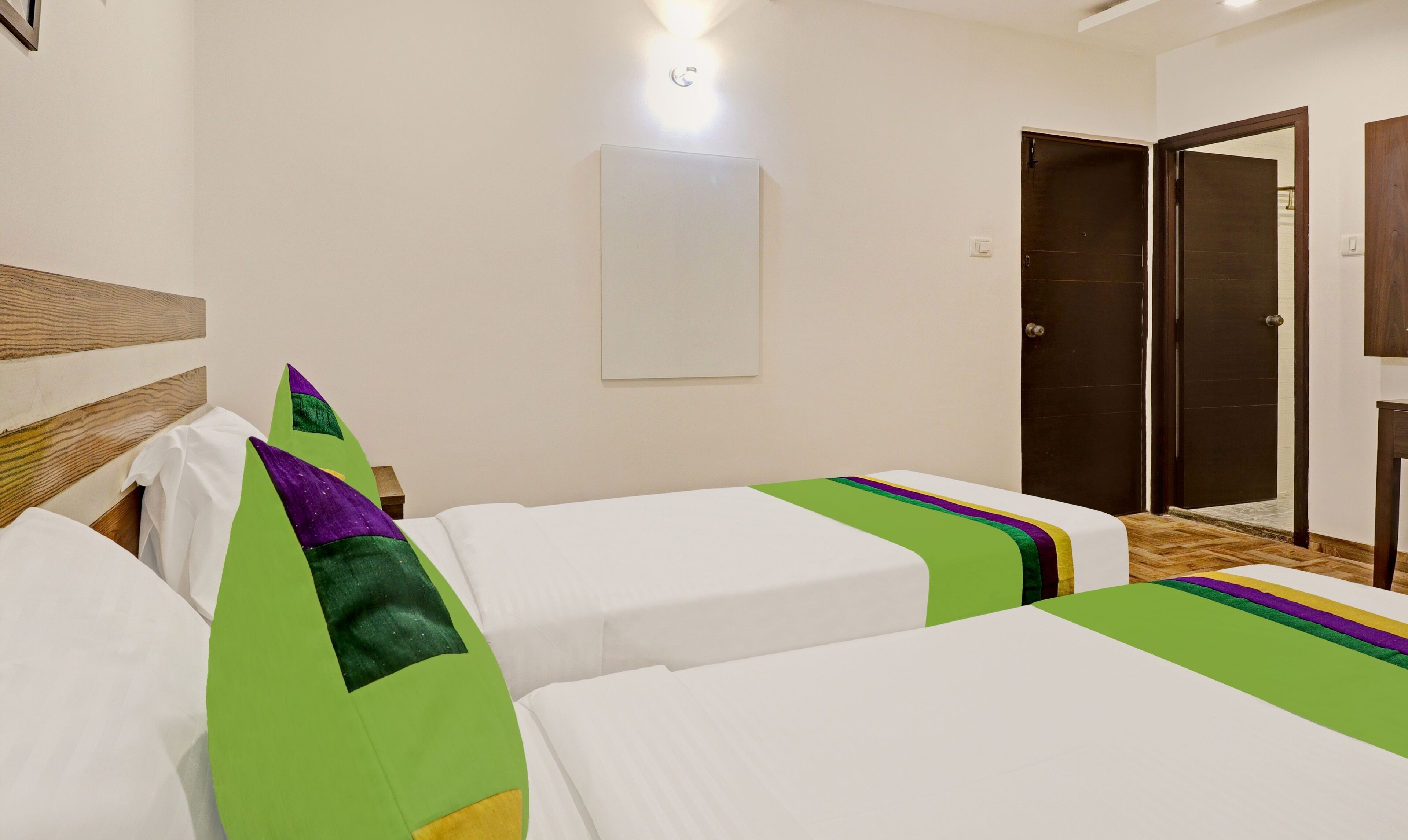 Habitación de hotel con dos camas individuales, cabecera de madera, almohada decorativa verde y morada, pared blanca, puerta y lámpara.