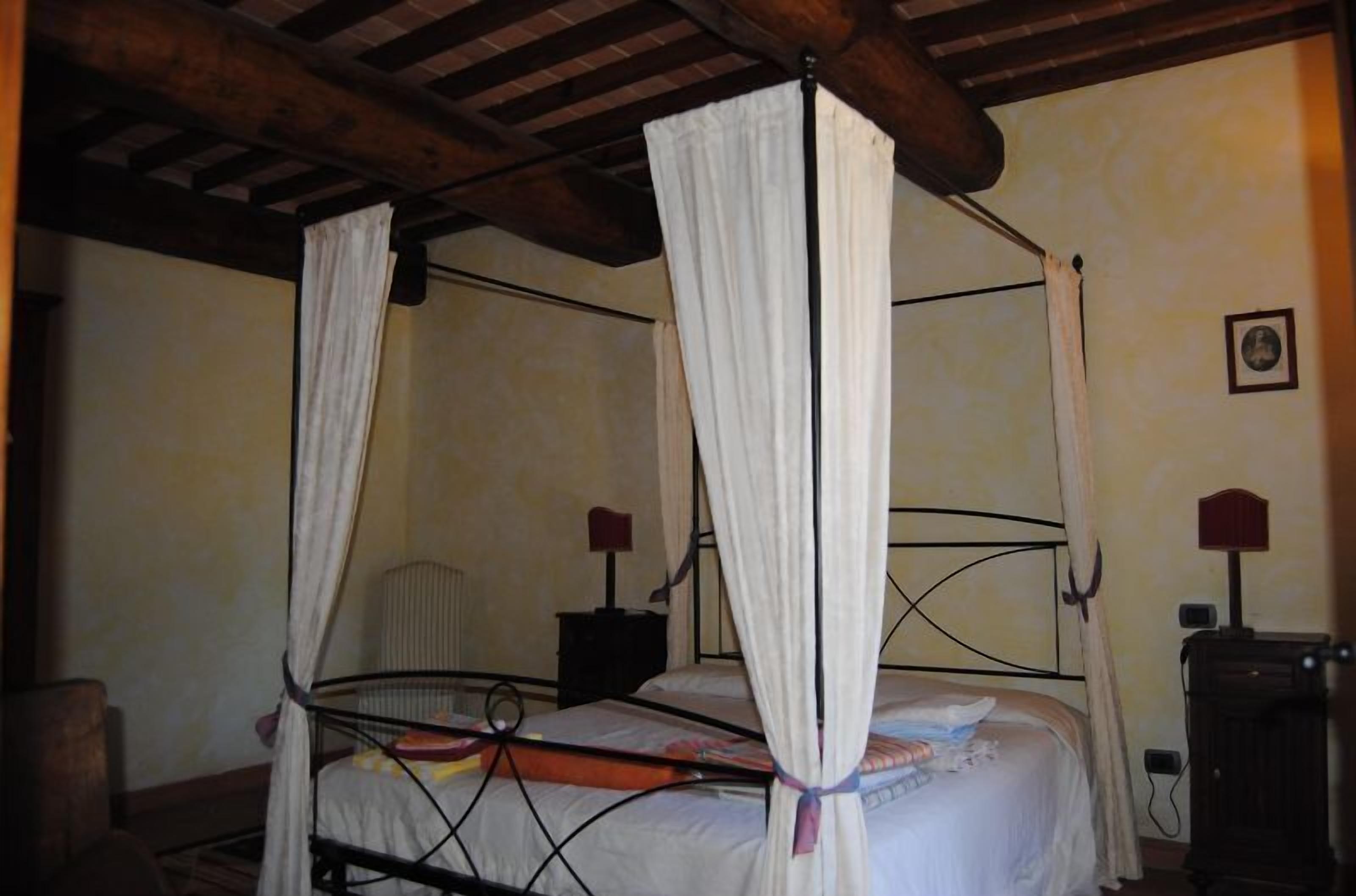 A four-poster bed with white curtains, a wooden ceiling, and a nightstand with a lamp.