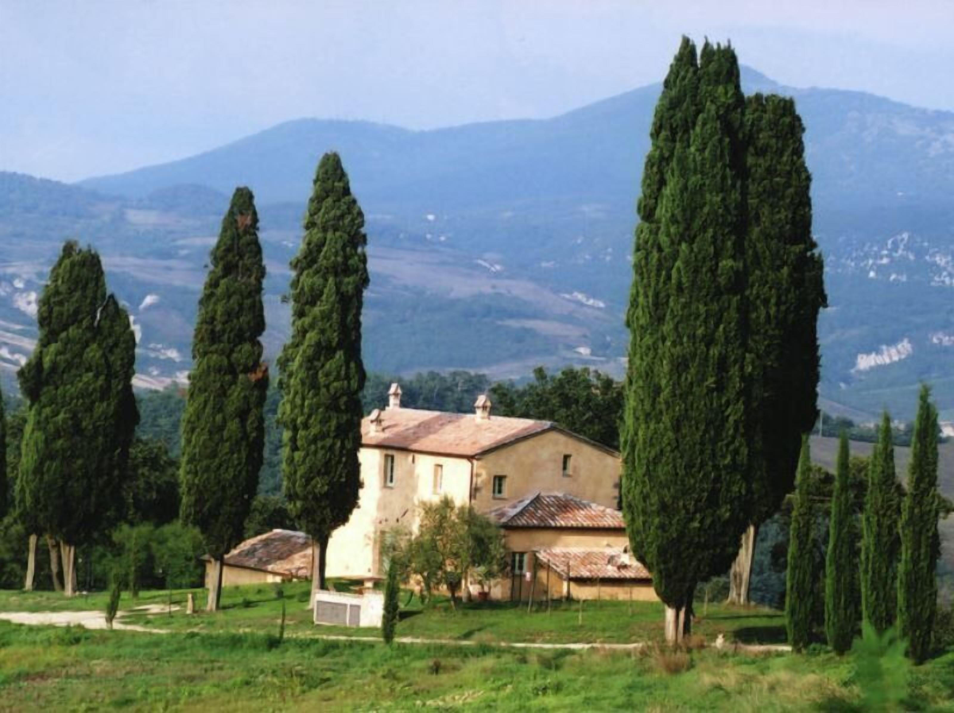 A rural house with a tiled roof, surrounded by tall cypress trees, set against a mountainous backdrop.