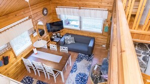 Interior de una cabaña de madera con comedor, sofá, televisor y ventanas.