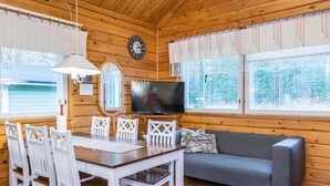 Un interior de cabaña acogedor, con paredes de madera, una mesa de comedor con sillas blancas, un sofá gris, un televisor de pantalla plana y ventanas grandes con cortinas.