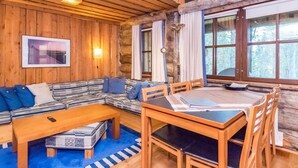 Interior acogedor de una cabaña de troncos con un sofá de madera, una mesa de comedor y un ventanal con vistas al bosque.