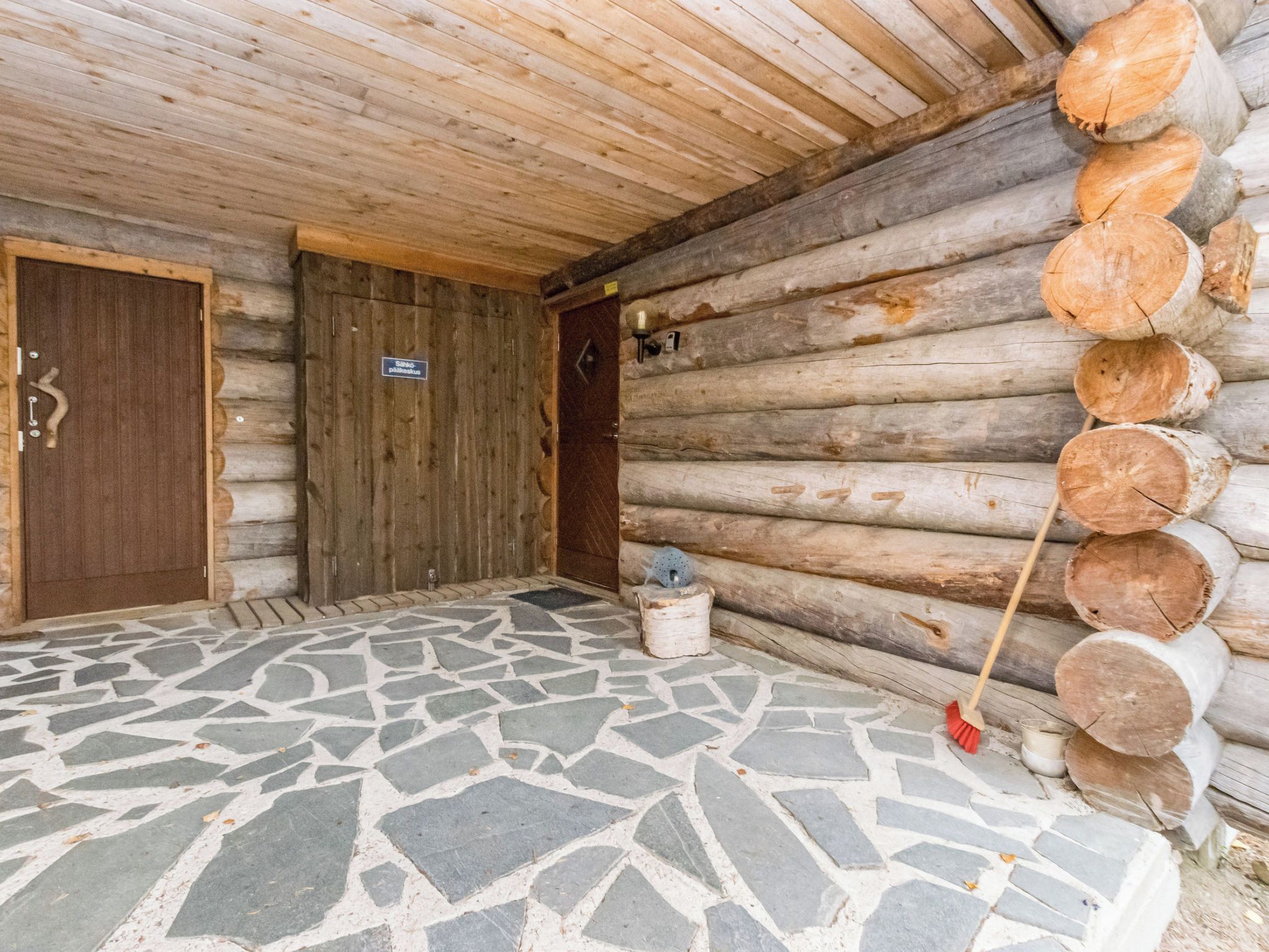 Eine Blockhütte mit einem Eingang aus Steinplatten, einer Holztür und einem Holzstoß an der Wand.