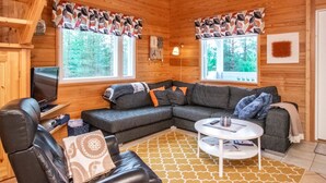 Un intérieur de chalet chaleureux avec un canapé d’angle, un fauteuil en cuir, une petite table ronde et un téléviseur.