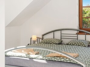 A bed with a geometric patterned bedspread, two pillows with a similar pattern, a bedside lamp, and a window with a view of greenery.