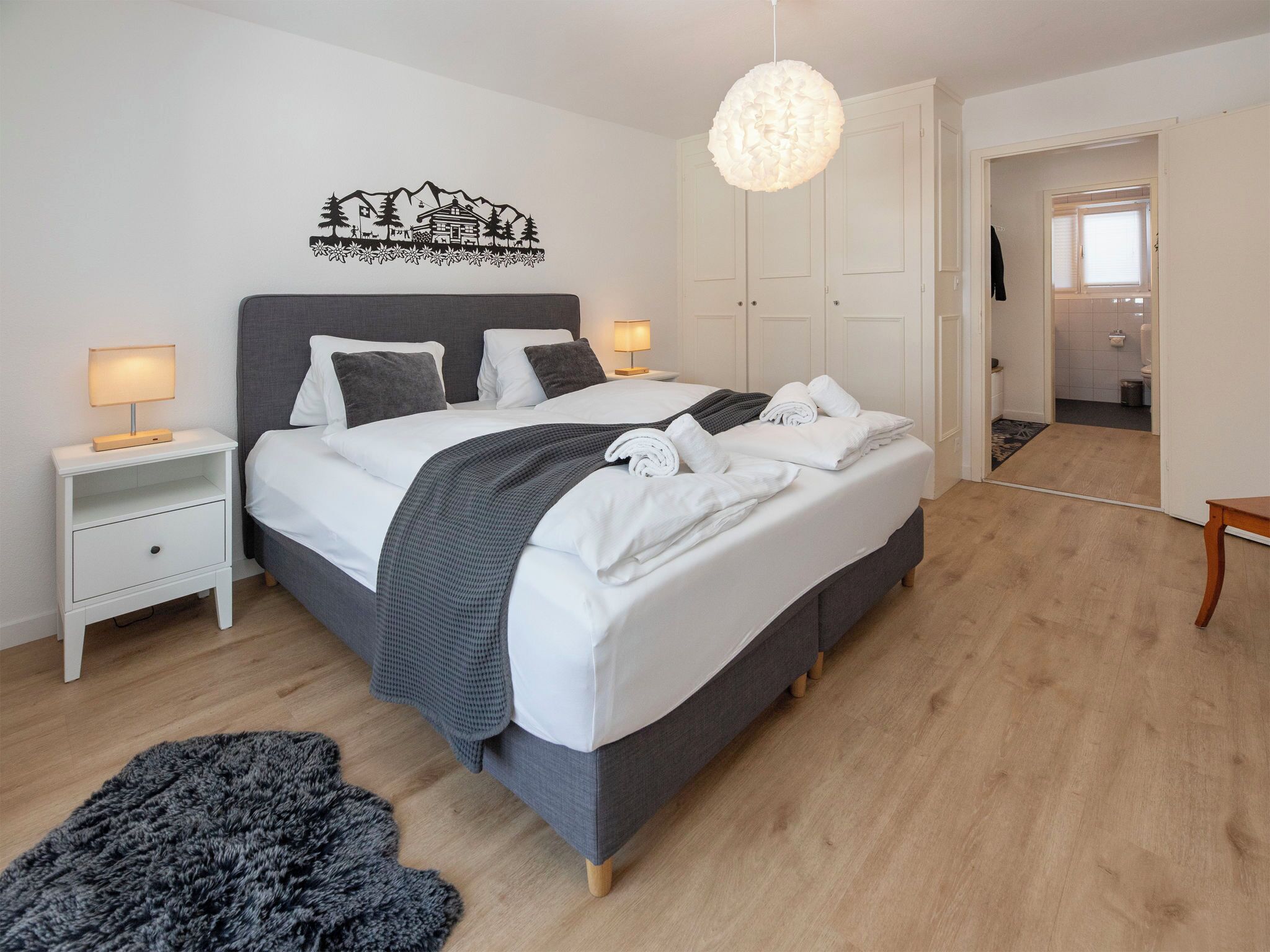 A neatly made bed with a grey headboard, white bedding, and a grey blanket. There are two bedside tables with lamps, a wall art piece, and a hanging light fixture.