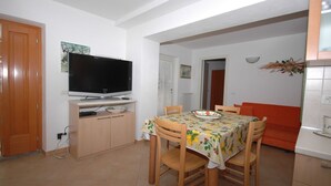 Un comedor con una televisión sobre un mueble bajo, una mesa con mantel floral y un sofá al fondo.