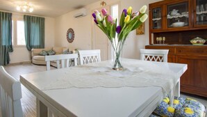 Un coin repas avec une table blanche, des chaises et un vase de tulipes. On y trouve également un buffet en bois contenant de la vaisselle, ainsi qu’une fenêtre avec des rideaux.