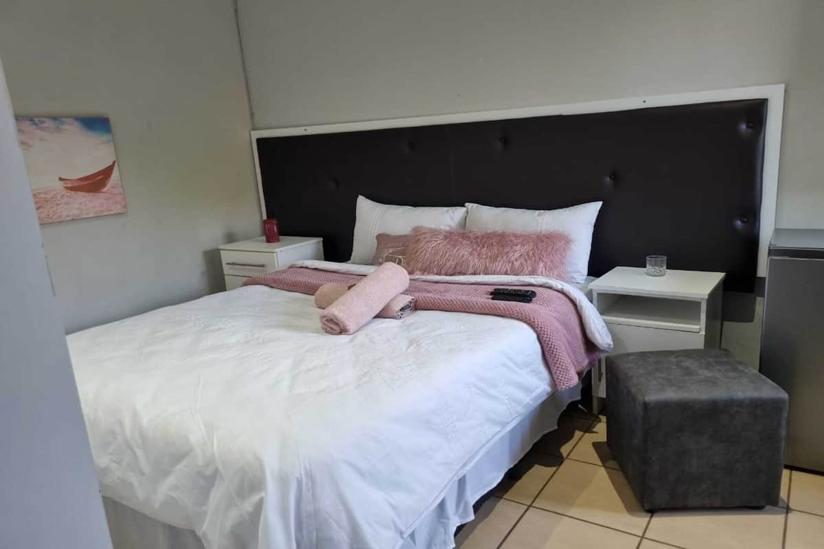 A neatly made bed with a white comforter and pink accents, a small bedside table with a glass, and a grey ottoman.