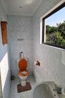 Un baño con inodoro, lavamanos y bañera. Una ventana ofrece vista a follaje exterior.