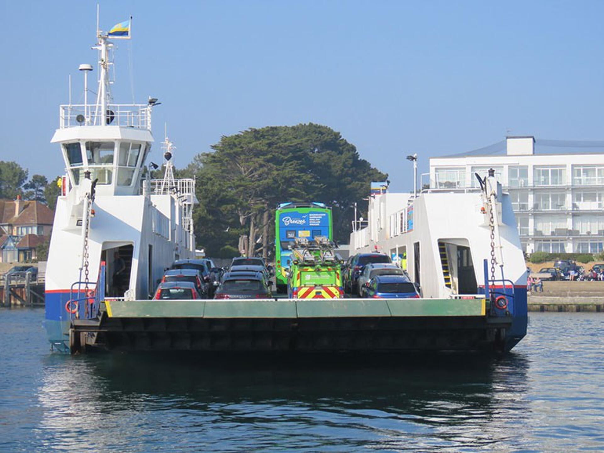 A ferry with multiple cars and a green truck on board, docked at a pier with buildings and trees in the background.