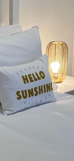 A bed with a white pillow that reads "HELLO SUNSHINE" and a wicker lamp with a light bulb.