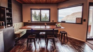 Une cuisine moderne avec un plafond en bois, de grandes fenêtres offrant une vue sur les arbres et les montagnes, un banc intégré et un coin repas avec des tabourets.